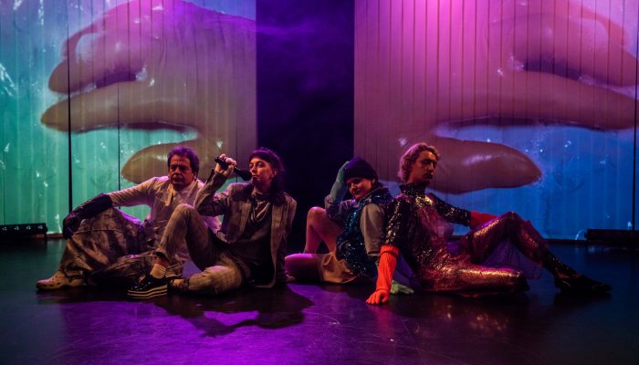 Stadttheater Vier Personen sitzen vor einer Leinwand mit zwei Händen in Großaufnahme. Eine Person hält ein Mikrofon in der Hand.