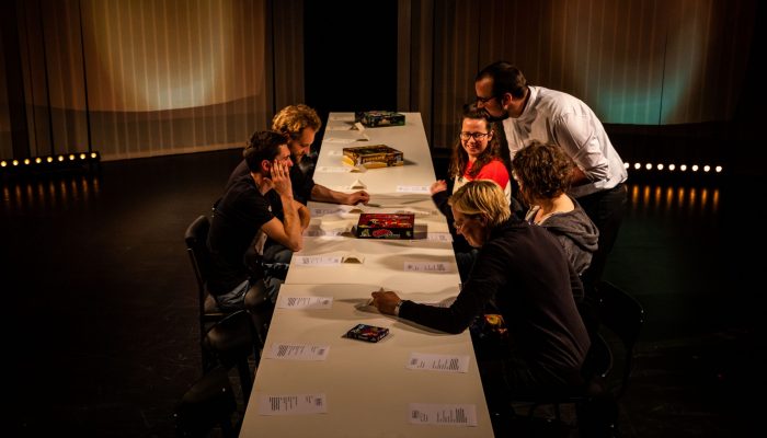 Stadttheater Fünf Menschen sitzen auf der Bühne an einem langen Tisch, Darauf Zettel und Spielekartons. Eine Person beugt sich zu ihnen und erklärt etwas.