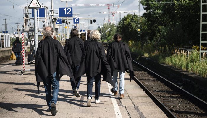 Rübermachen Fünf Menschen mit schwarzen Umhängen gehen auf einem Bahnsteig von der Kamera weg. Gezeigt wird ihre Rückansicht.