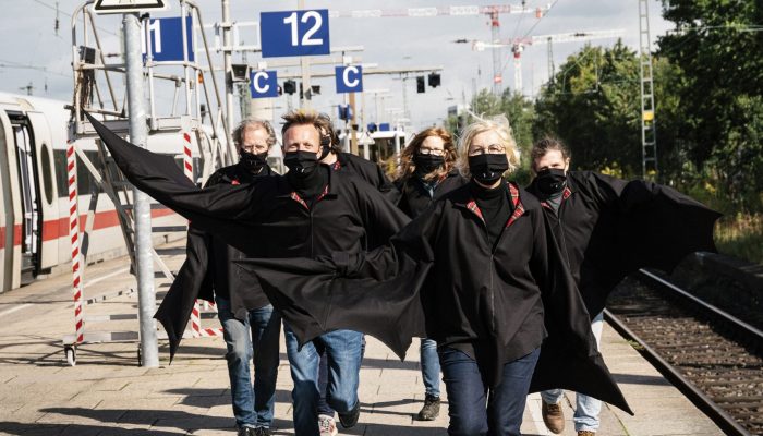 Rübermachen Sechs als Fledermaus verkleidete Menschen laufen mit zum Teil ausgebreiteten Armen auf einem Bahnsteig auf die Kamera zu.