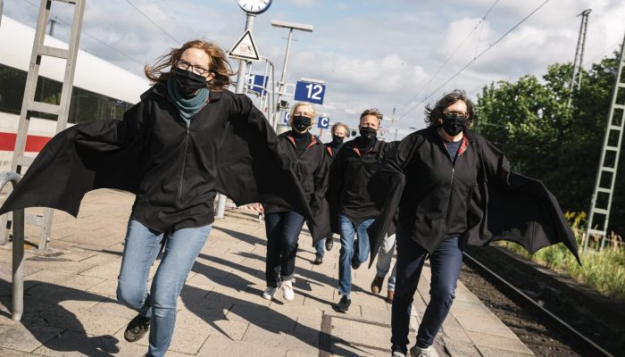 Rübermachen Eine Gruppe als Fledermaus verkleideter Menschen rennt auf einem Bahnsteig auf die Kamera zu. Die Arme sind weit ausgebreitet.