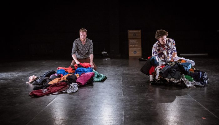 Stadttheater Zwei Menschen hantieren mit Kleidungsstücken, die jeweils in einem großen Haufen vor ihnen liegen.