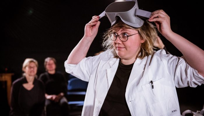 Stadttheater Eine Frau mit weißem Kittel setzt sich eine Schutzbrille auf. Im Hintergrund ist das Publikum zu sehen.