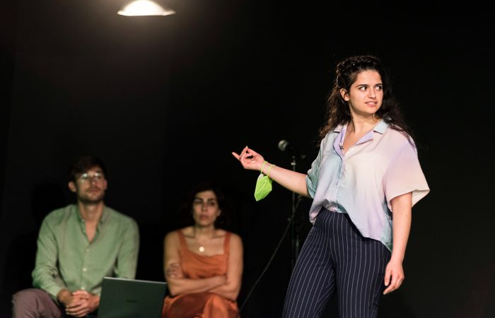 citizenpark_1 Eine junge, nach links schauende Frau streckt den rechten Arm aus, an dem ein Mund-Nasenschutz hängt. Im Hintergrund sitzen ein Mann und eine Frau.