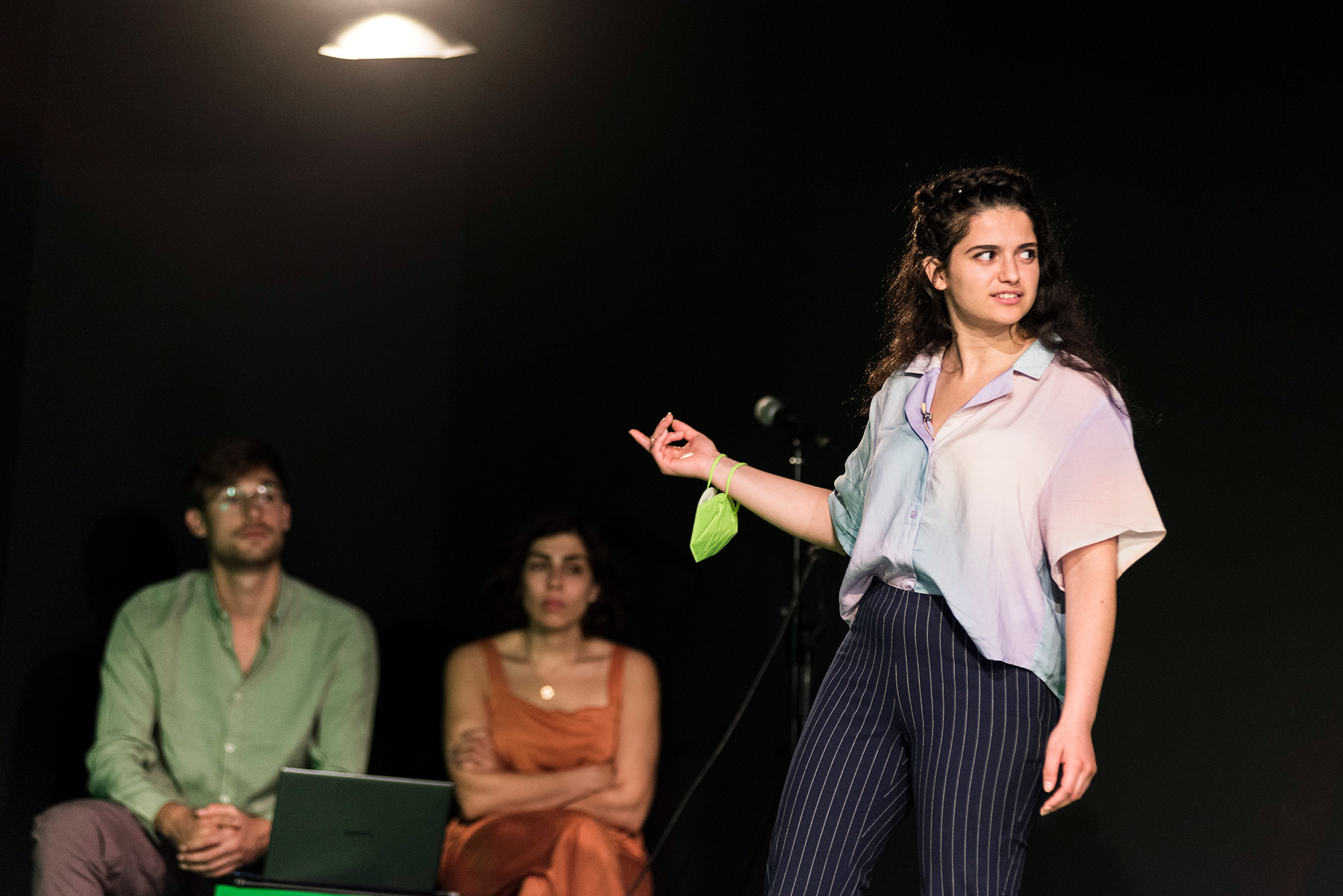 Eine junge, nach links schauende Frau streckt den rechten Arm aus, an dem ein Mund-Nasenschutz hängt. Im Hintergrund sitzen ein Mann und eine Frau.