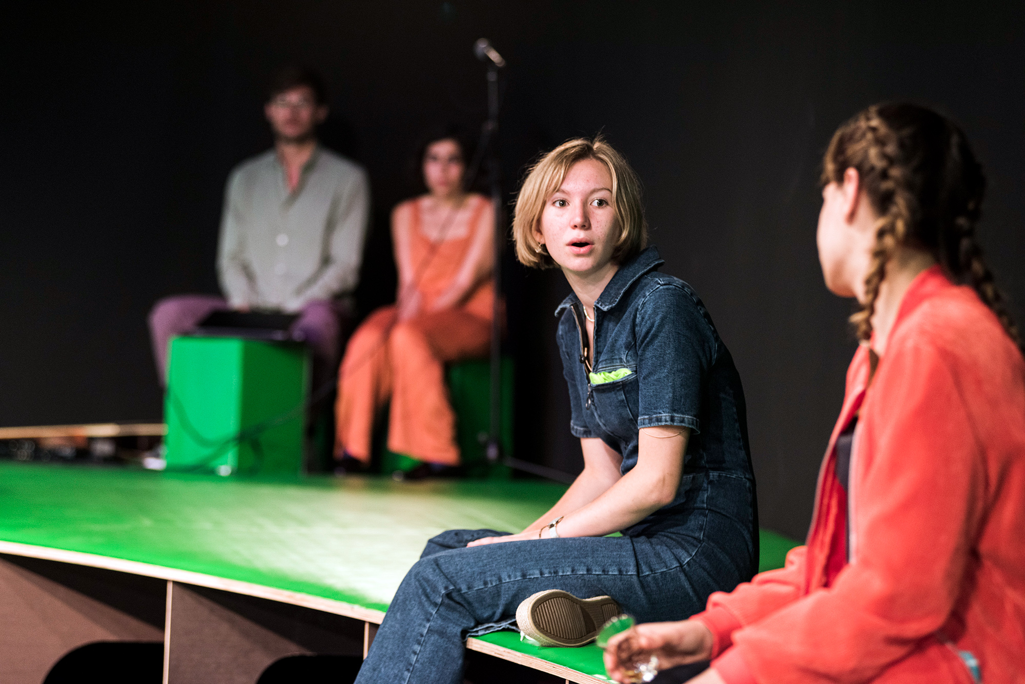 Zwei junge, weiblich gelesene Personen unterhalten sich. Im Hintergrund sitzen zwei weitere Personen und hören zu.