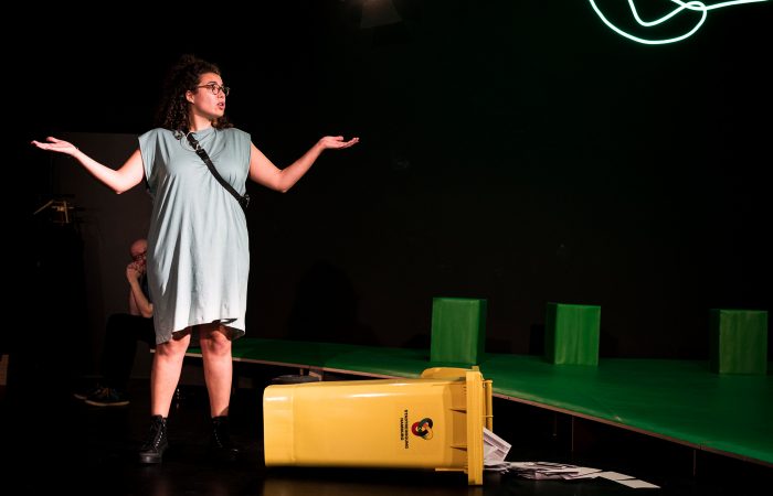 citizenpark_7 Eine junge Person im Kleid hebt achselzuckend die Arme. Sie steht vor einer umgekippten gelben Recyclingtonne.