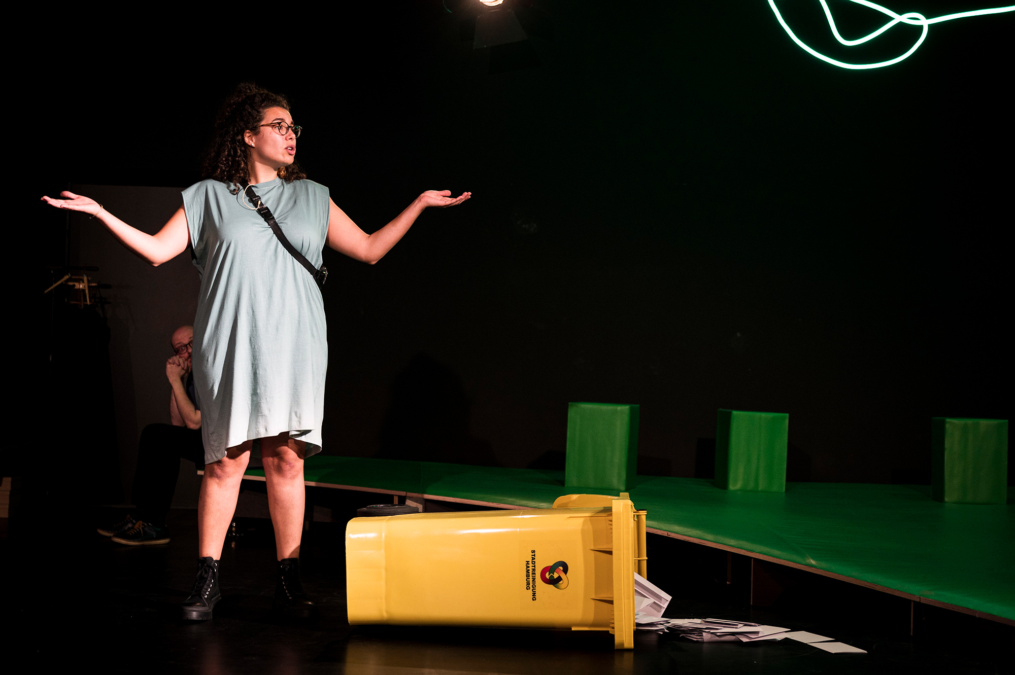 Eine junge Person im Kleid hebt achselzuckend die Arme. Sie steht vor einer umgekippten gelben Recyclingtonne.