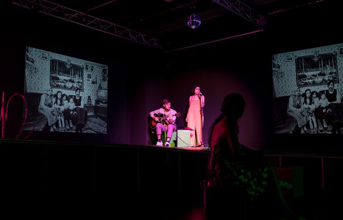 citizenpark_5 Ein dunkler Raum. Rechts und links zwei Leinwände mit schwarz-weißen Familienfotos. In der Mitte ein Gitarrist und eine Sängerin im Scheinwerferlicht..