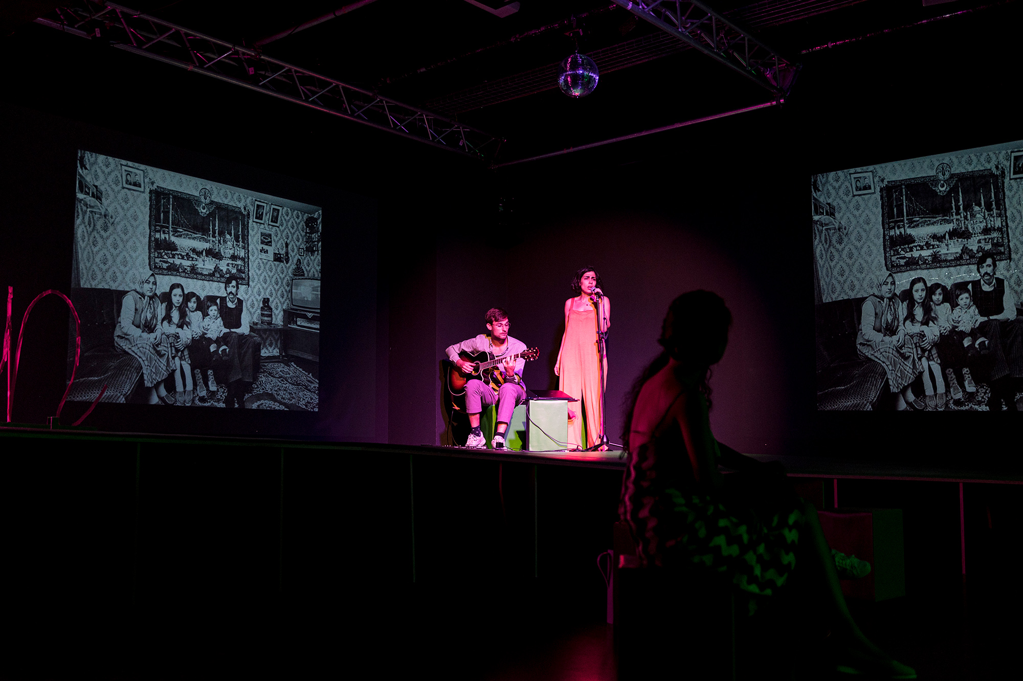 Ein dunkler Raum. Rechts und links zwei Leinwände mit schwarz-weißen Familienfotos. In der Mitte ein Gitarrist und eine Sängerin im Scheinwerferlicht..