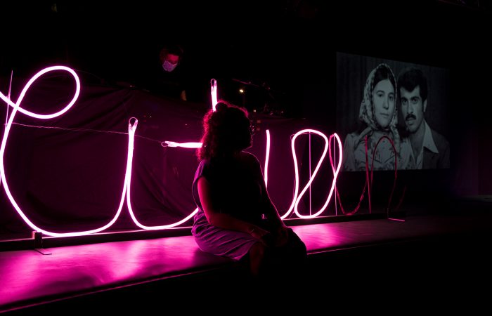 citizenpark_8 Eine Person sitzt im Dunklen vor einer rosafarbenen Leuchtschrift. Rechts im Hintergrund ist ein Schwarz-weiß-Foto eines Migrantenpaares eingeblendet.