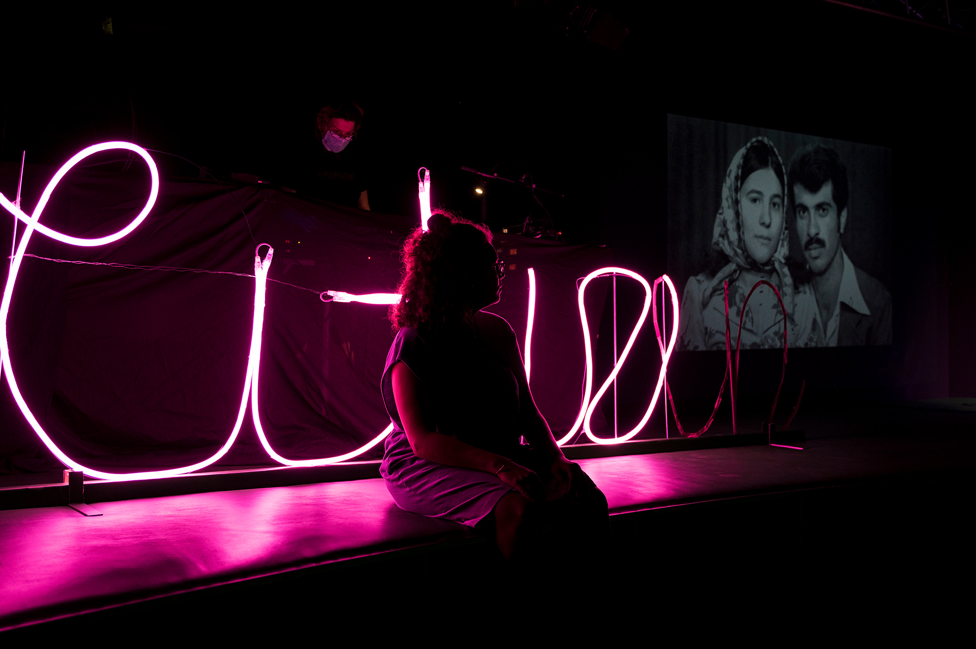 Eine Person sitzt im Dunklen vor einer rosafarbenen Leuchtschrift. Rechts im Hintergrund ist ein Schwarz-weiß-Foto eines Migrantenpaares eingeblendet.