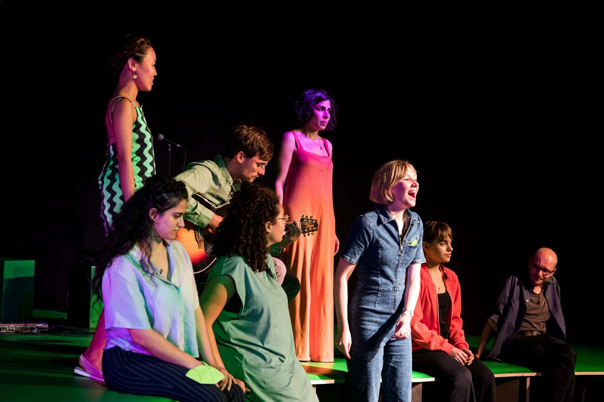 Sieben junge Menschen stehen um einen sitzenden Gitarristen und singen ein Lied.