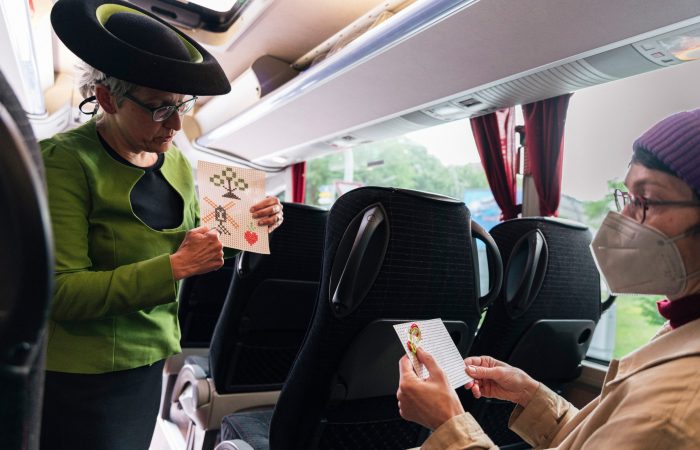 1 Gutes aus Hamburger Landen © Isabel Machado Rios Eine Szene im Bus: eine Frau im grünen Kostüm mit Hut erklärt einer Mitfahrerin ein Stickmuster.