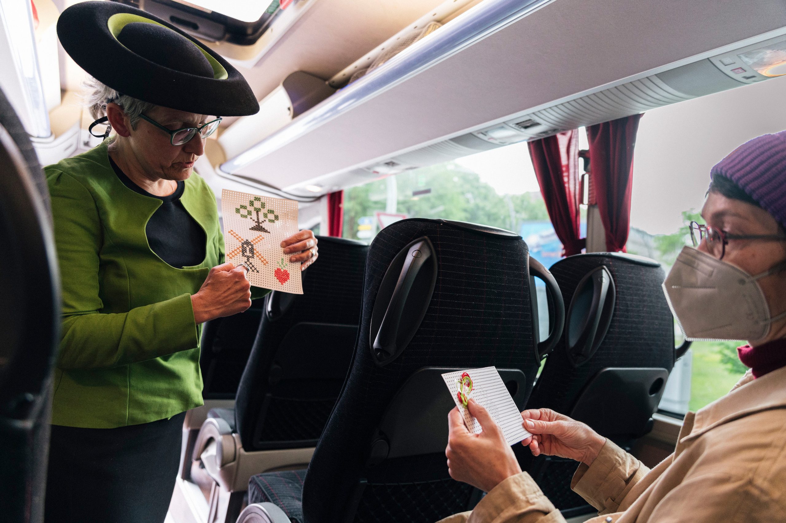Eine Szene im Bus: eine Frau im grünen Kostüm mit Hut erklärt einer Mitfahrerin ein Stickmuster.