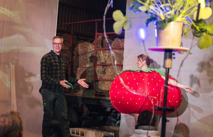 4 Gutes aus Hamburger Landen © Isabel Machado Rios Ein Mann im Flanellhemd zeigt auf eine als Erdbeere verkleidete, tanzende Frau. Im Vordergrund ist eine Pflanze mit Bewässerungsschläuchen.