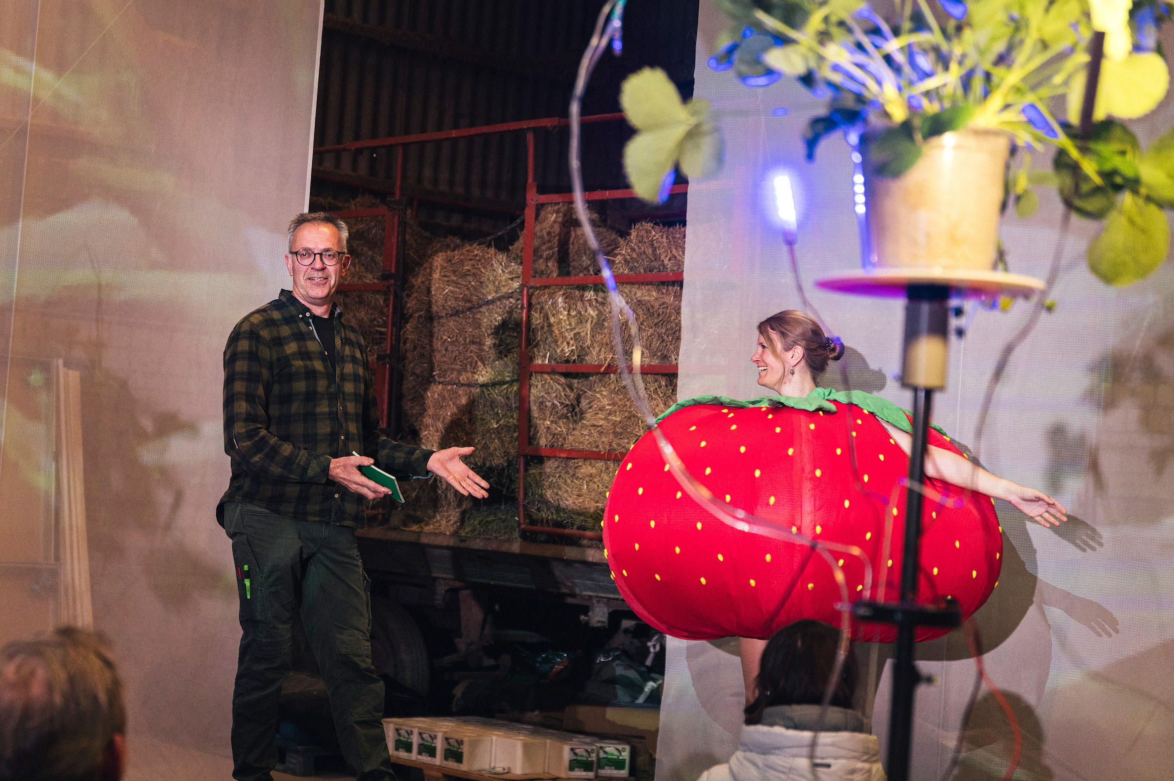 Ein Mann im Flanellhemd zeigt auf eine als Erdbeere verkleidete, tanzende Frau. Im Vordergrund ist eine Pflanze mit Bewässerungsschläuchen.