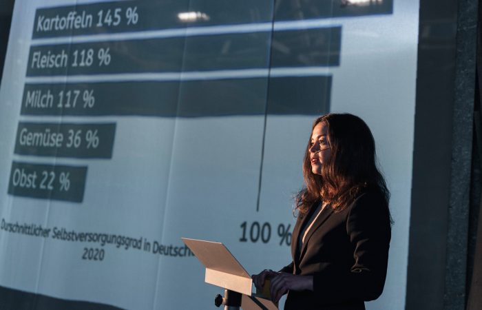 5 Gutes aus Hamburger Landen © Isabel Machado Rios Eine junge Frau hält einen Vortrag vor einer Leinwand mit statistischen Zahlen.