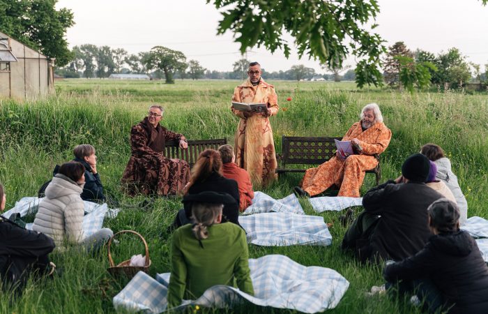6 Gutes aus Hamburger Landen © Isabel Machado Rios.jpg Auf einer Wiese drei Männer in braunen Roben. Zwei sitzen, einer steht und hält einen Vortrag. Im Vordergrund sitzen die Zuhörenden auf Picknickdecken.