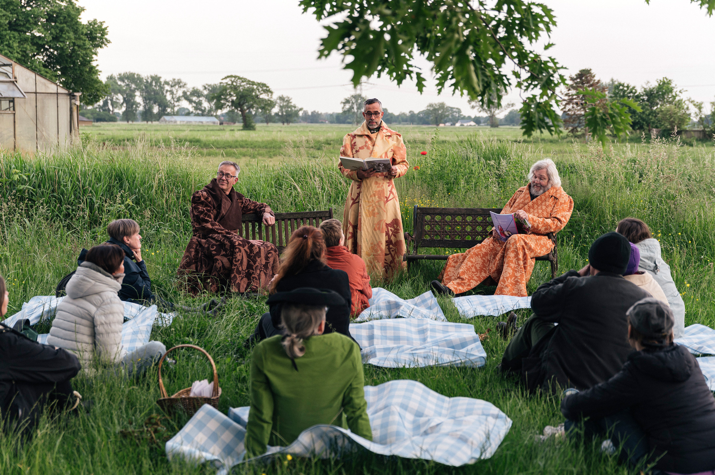 Auf einer Wiese drei Männer in braunen Roben. Zwei sitzen, einer steht und hält einen Vortrag. Im Vordergrund sitzen die Zuhörenden auf Picknickdecken.