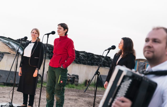 8 Gutes aus Hamburger Landen © Isabel Machado Rios.jpg.jpg Drei junge Frauen stehen auf einem Bauernhof vor drei Mikrophonen. Im Vordergrund. unscharf ein Mann mit Akkordeon.