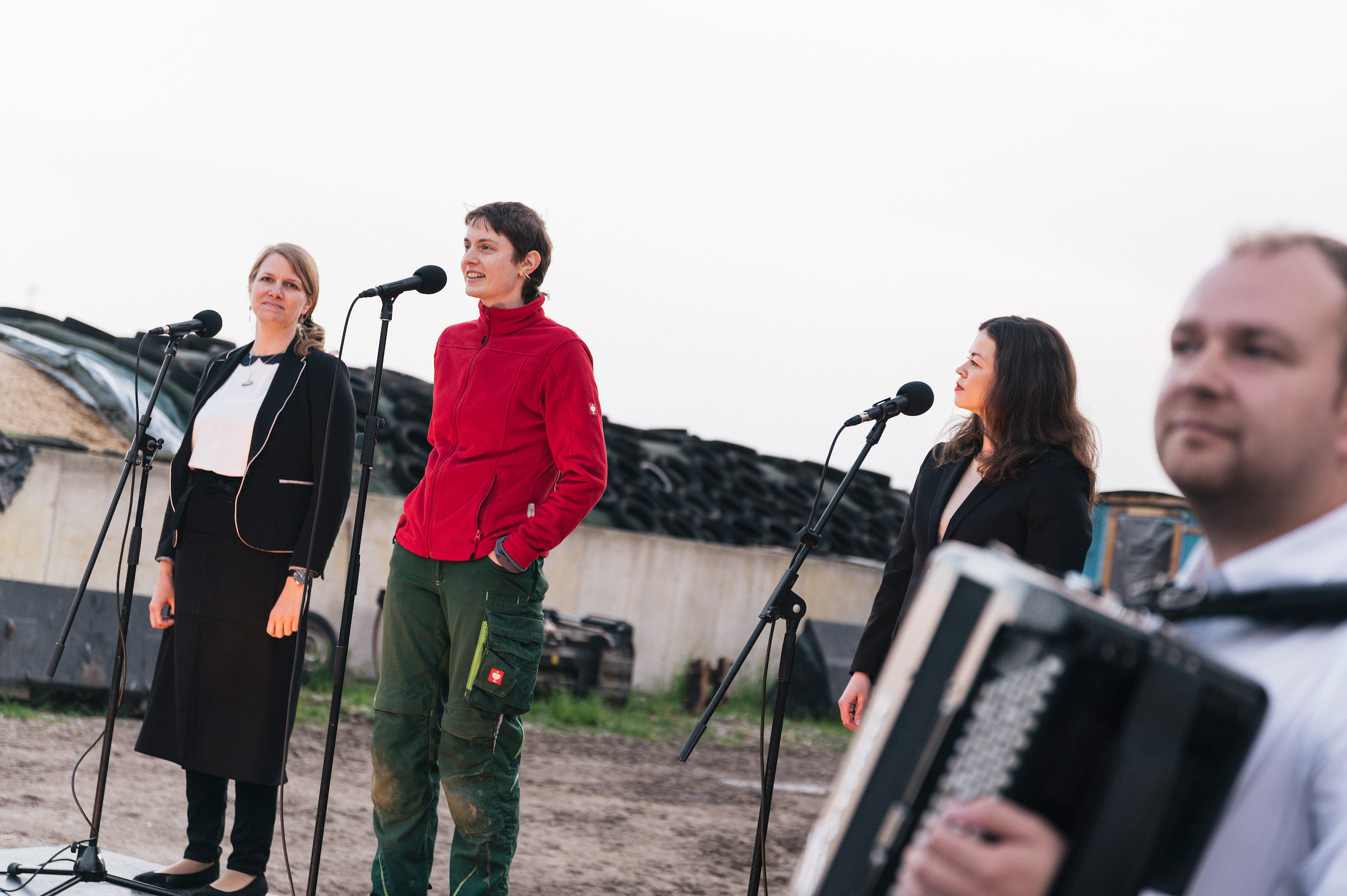 Drei junge Frauen stehen auf einem Bauernhof vor drei Mikrophonen. Im Vordergrund. unscharf ein Mann mit Akkordeon.