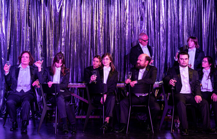 JudenJudenJuden-IsabelMachadoRios-4360_web Neun Menschen im Frack sitzen an einem langen Tisch und haben Sektgläser in der Hand.. Im Hintergrund ist ein glänzender Vorhang.