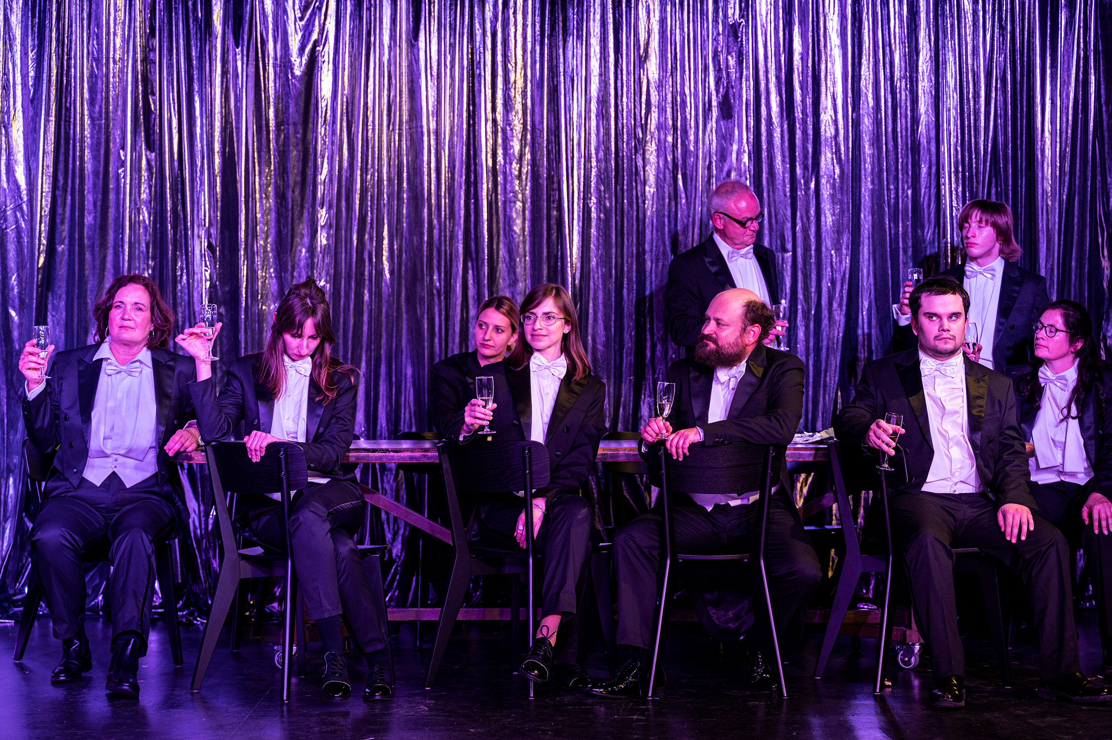 Neun Menschen im Frack sitzen an einem langen Tisch und haben Sektgläser in der Hand.. Im Hintergrund ist ein glänzender Vorhang.