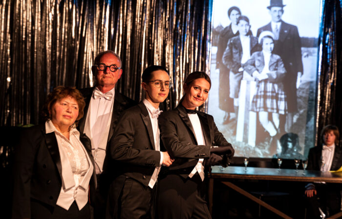JudenJudenJuden-IsabelMachadoRios-4446_web Vier Personen im Frack sthen an einem Tisch mit Sektgläsern vor einer Leinwand mit alten Familienfotos.