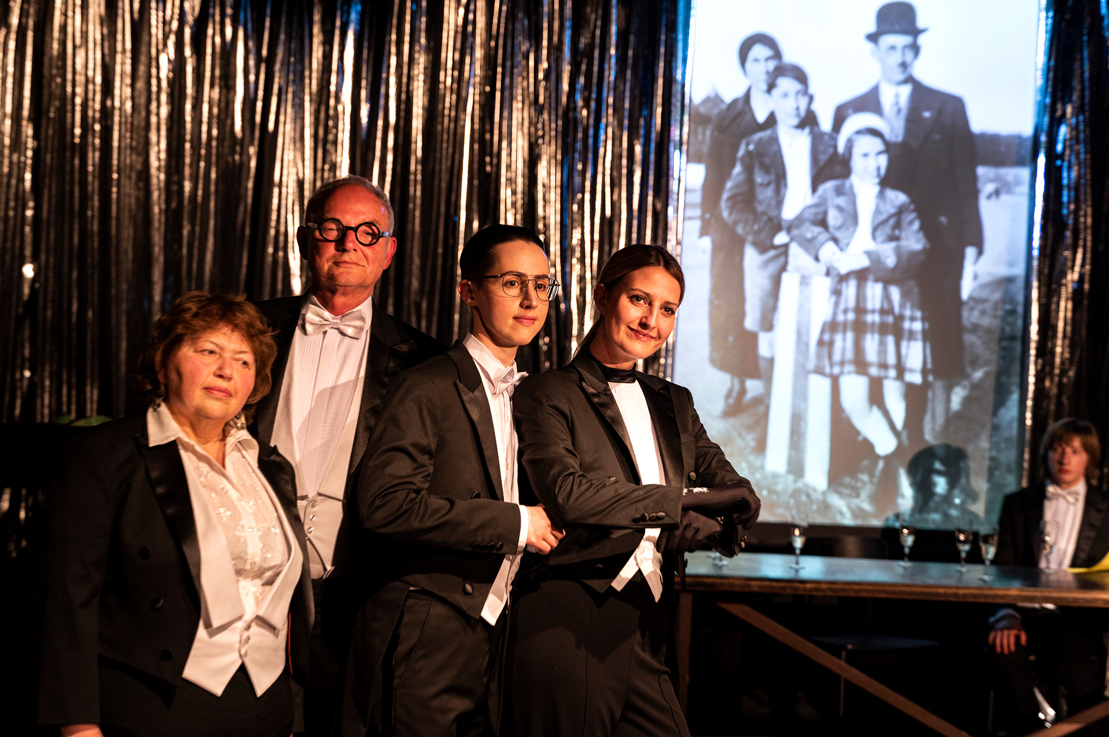 Vier Personen im Frack sthen an einem Tisch mit Sektgläsern vor einer Leinwand mit alten Familienfotos.