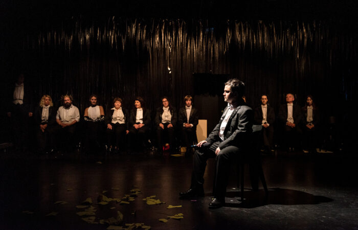 JudenJudenJuden-IsabelMachadoRios-7390_web Eine Person sitzt im Rampenlicht auf einem Stuhl, ein Mikrofon in der Hand. Im Hintergrund sitzen elf Menschen und hören zu.