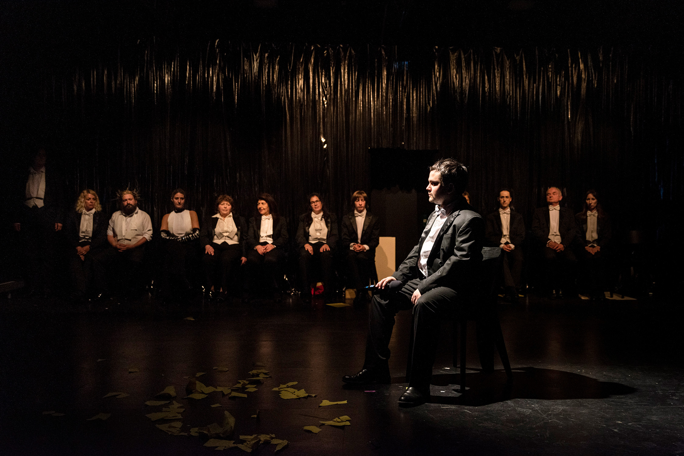 Eine Person sitzt im Rampenlicht auf einem Stuhl, ein Mikrofon in der Hand. Im Hintergrund sitzen elf Menschen und hören zu.