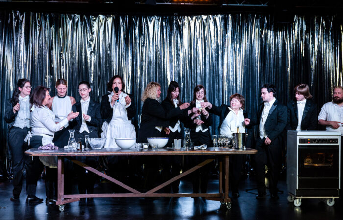 JudenJudenJuden-IsabelMachadoRios-7424_web Zwölf festlich gekleidete Personen stehen hinter einem Tisch. Darauf Schüsseln, Sektkühler und Sektgläser.