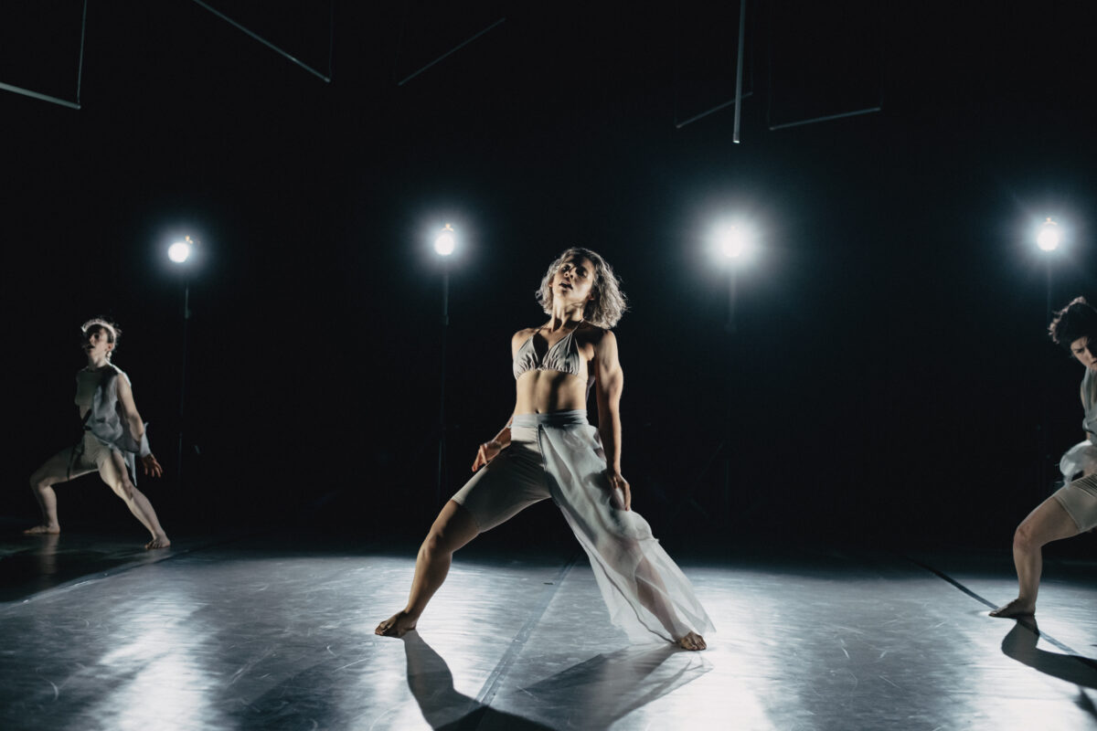 Drei junge Tänzerinnen auf einer Bühne, Eine Frau ist in der Mitte, schaut nach oben und steht in einem breiten Schritt.