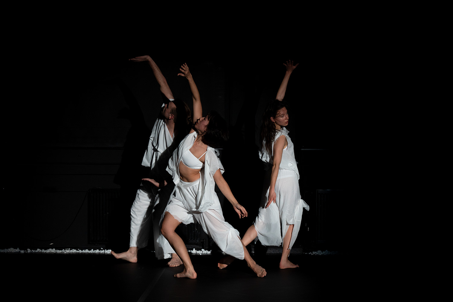 Drei Tänzerinnen in weißer Kleidung vor schwarzem Hintergrund stehen zusammen und machen einen Ausfallschritt, ein Arm ist hoch nach oben gestreckt, der andere zeigt nach unten