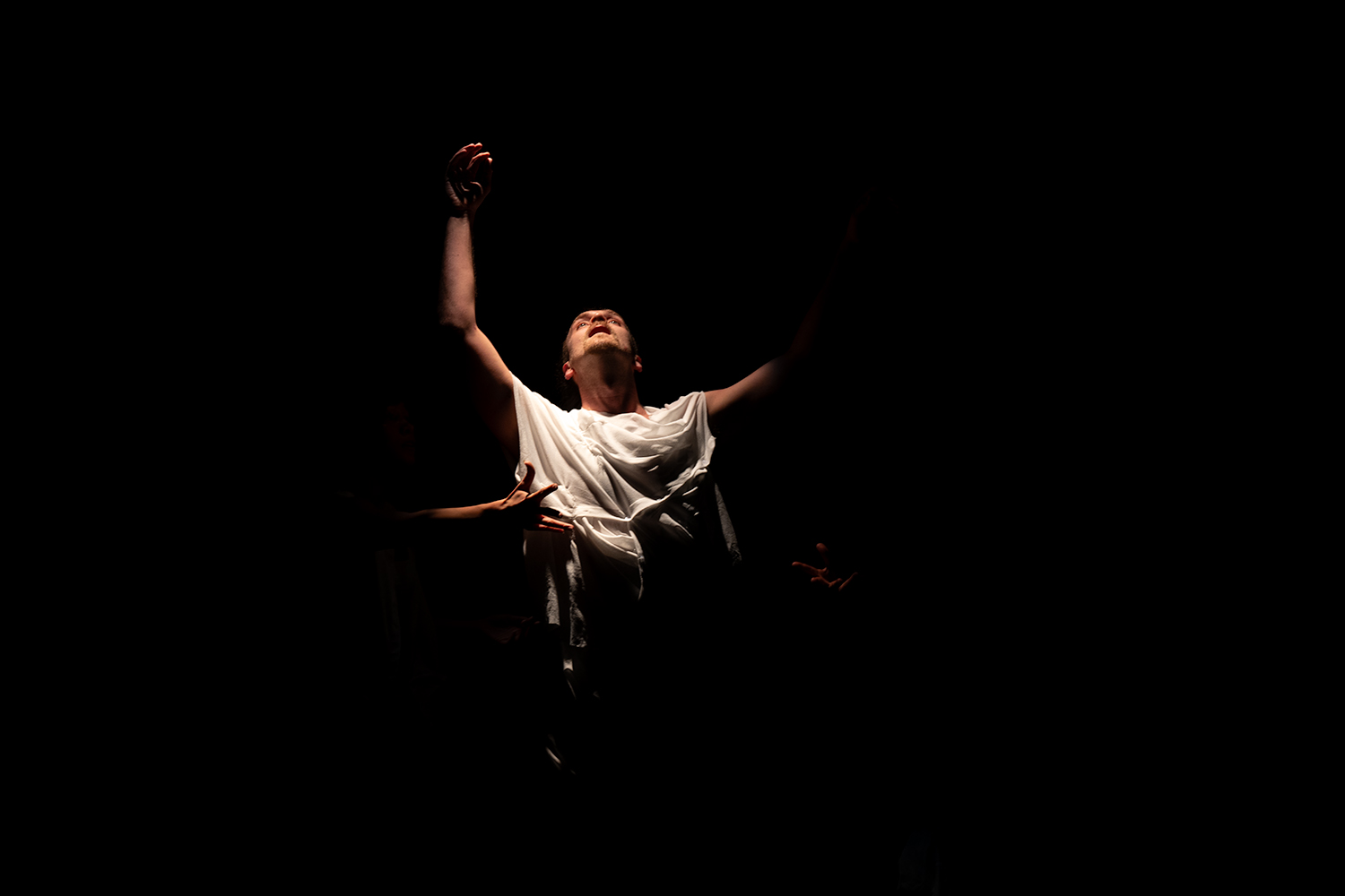 In der Mitte eines dunklen Raumes steht ein Tänzer im Lichtkegel, schaut nach oben und streckt die Hände in die Luft. von den Seiten her ragen zwei weitere Hände ins Bild.