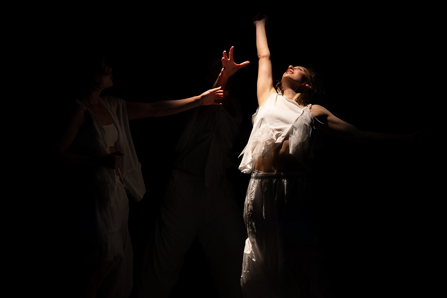 in einem dunklen Raumes steht ein Tänzerin im Lichtkegel, sie schaut nach oben und streckt den rechten Arm nach oben. von rechts her ragen weitere Hände ins Bild.