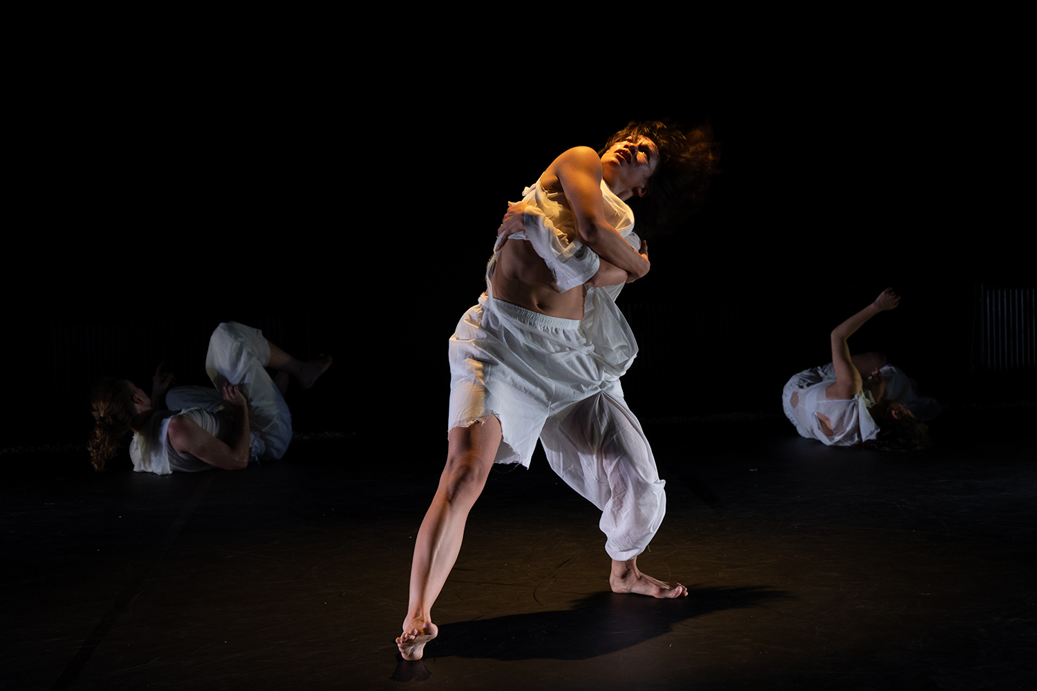 Im Hintergrund rollen sich zwei Tänzer:innen am Boden, im Vordergrund ist eine Tänzerin mit verschränkten Armen, das Gesicht nach oben gerichtet.