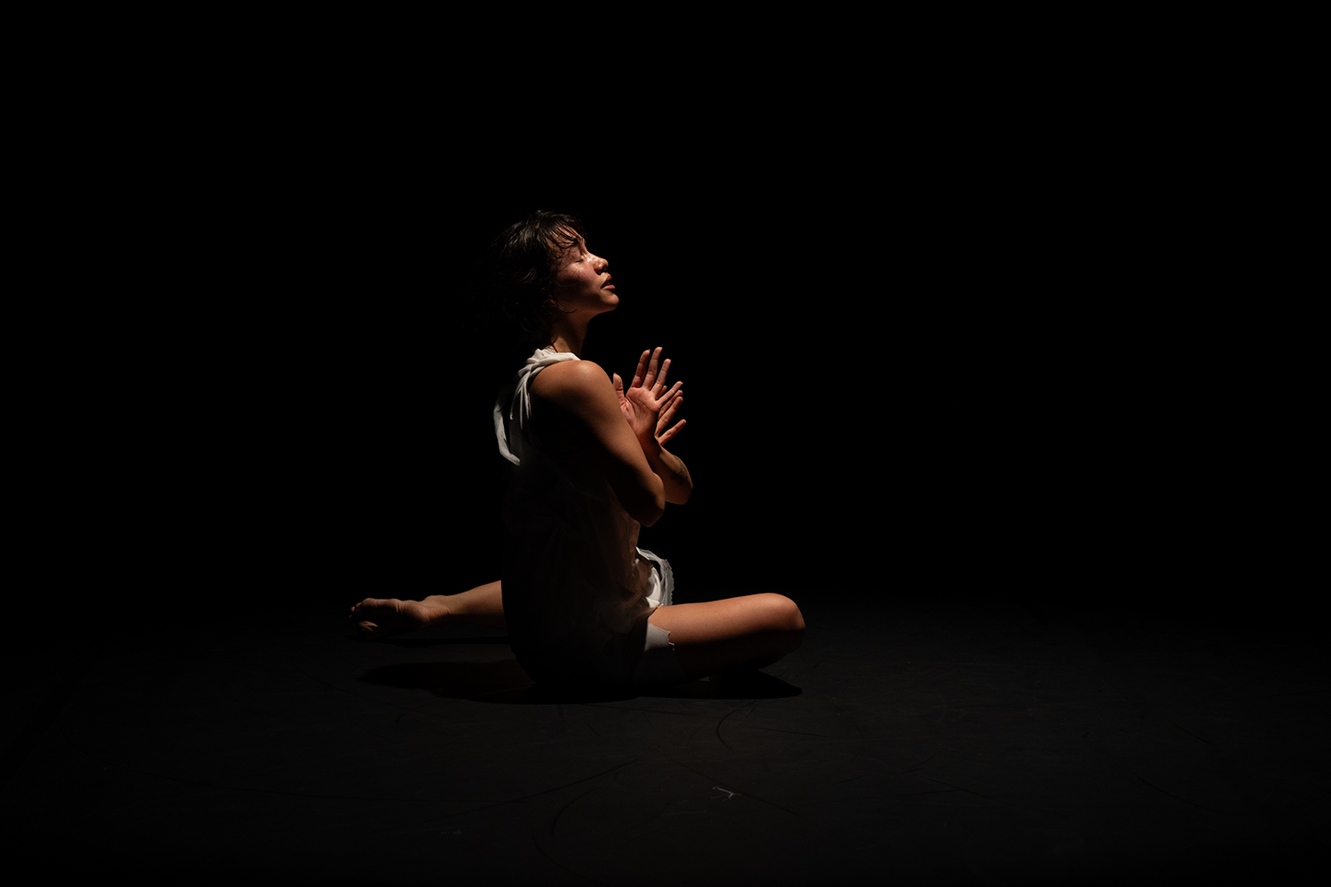 In einem dunklen Raum sitz eine Tänzerin mit geschlossenen Augen am Boden, verschränkt die Arme mit weit aufgefächerten Händen vor der Brust