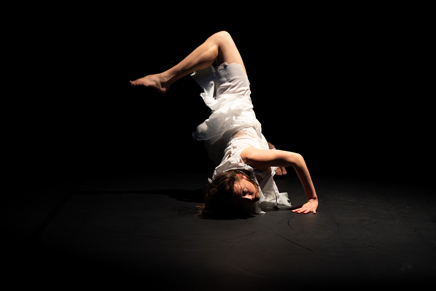 Eine in weiß gekleidete Tänzerin vor schwarzem Hintergrund in einer verdrehten Pose, Kopf und Arm am Boden abgestützt, das linke angewinkelte Knie nach hinten verdreht.