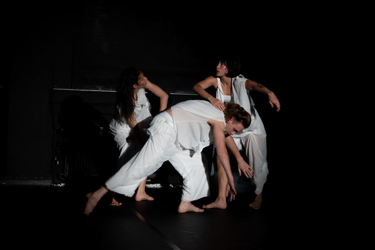 In einem dunklen Raum bewegen sich drei Tänzer:innen. In der Mitte ein Tänzer mit großem Ausfallschritt, vornübergebeugt, die Arme zeigen zum Boden. Von links und rechts sind zwei Tänzerinnen über ihn gebeugt.