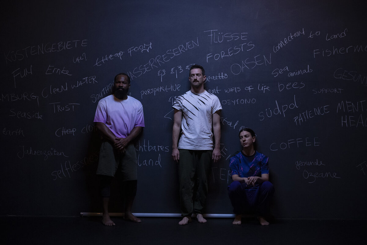 Zwei Tänzer und eine Tänzerin stehen vor einer schwarzen Wand, auf der mit Kreide Wörter wie "Perspektive", "Oasen" oder "Vertrauen" geschrieben steht.