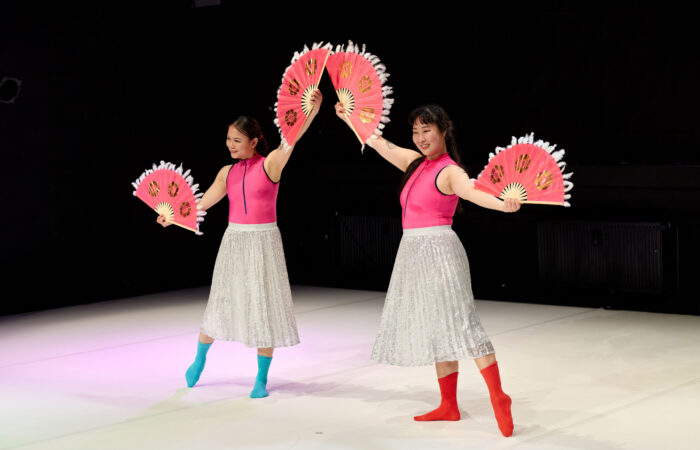 WE PRESENT Festival 2025 - Id Your Girlfriends Szenenfoto. zwei weiblich gelesene Personen posieren mit ausgestreckten Armen und Fächern in ihren Händen.nd.