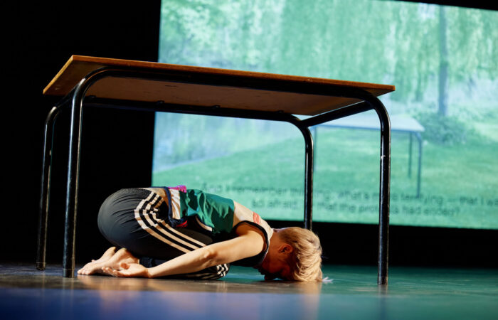 WE PRESENT Festival 2025 - No I can't forget tomorrow Szenenfoto: Eine Person kniet unter einem Tisch, der vor einer Leinwand steht. Ihr Kinn berührt den Boden. Die Hände sind nach hinten ausgestreckt.