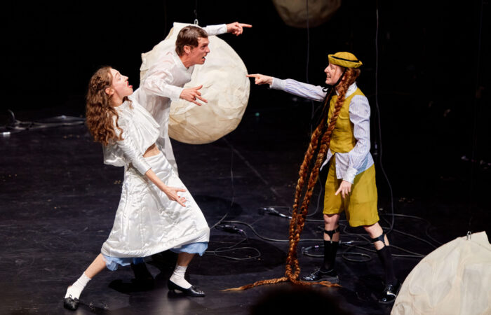 WE PRESENT Festival 2025 - Sandmann Szenenfoto: Eine Person mit bodenlangen Zöpfen steht, wie in einem Streit mit weit ausgestrecktem Arm, einem Mann und einer Frau gegenüber.