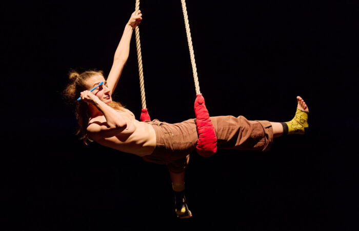 WE PRESENT Festival 2025 - geWOHNheiten Szenenfoto: Eine Frau hängt vertikal ausgestreckt in einem Hochtrapez und streicht mit einer Zahnbürste über eine ihrer Augenbrauen.