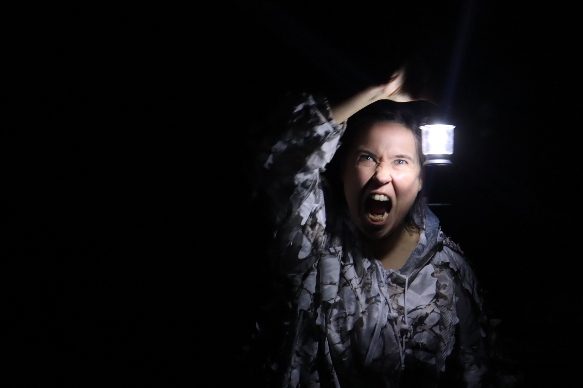 Eine Frau im Dunklen, angestrahlt nur von einer Campinglampüe in ihrer Hand. Das Gesicht zu einer brüllenden Fratze verzerrt.