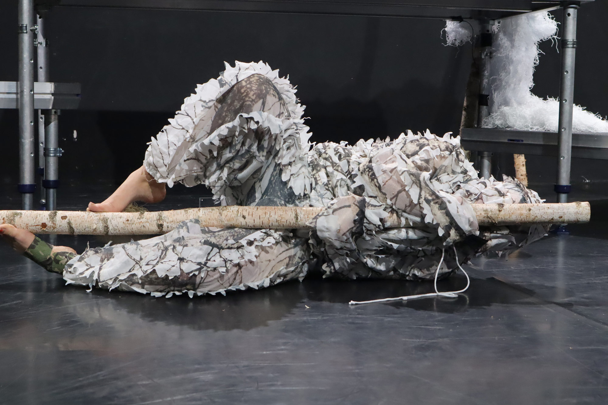 Eine Frau in weißem Tarnanzug umschlingt, auf dem Boden liegend, einen Baumast.